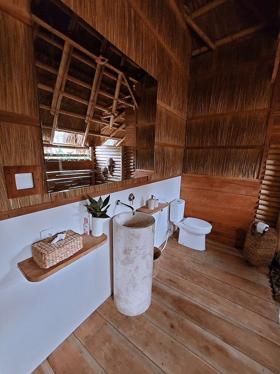 A bathroom featuring natural bamboo and wood elements, with a freestanding marble sink surrounded by wooden flooring. A mirror with a bamboo frame reflects the space, while a small potted plant adds a touch of greenery. Natural light filters through window openings.