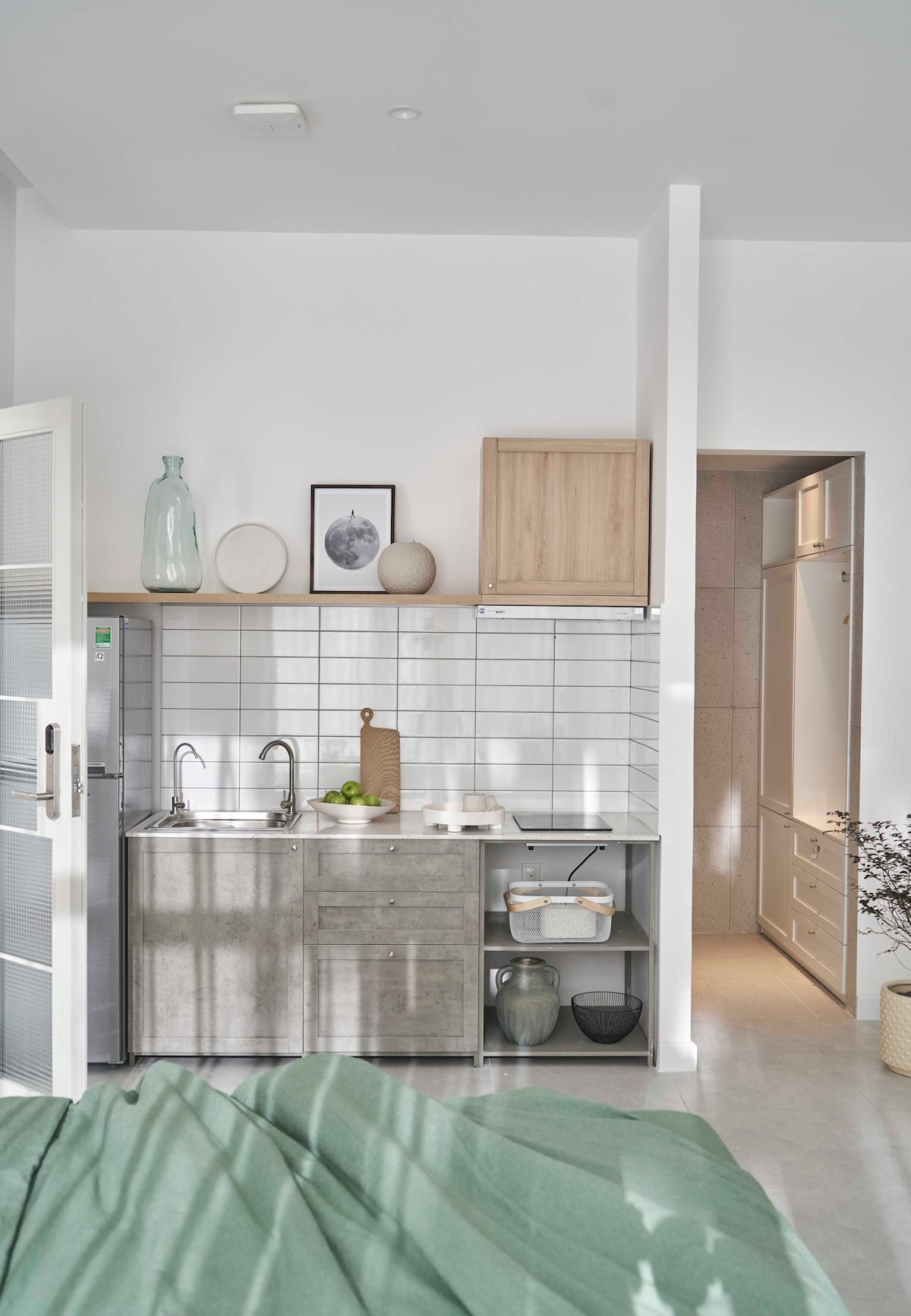A compact kitchen area features a stainless steel sink, minimalist cabinetry in light wood and gray tones, and white tiled backsplashes. Natural light illuminates the space, highlighting decorative items on the counter. A doorway leads to a separate room, enhancing the kitchen's functionality.