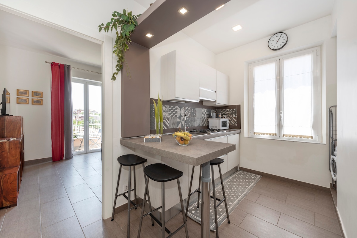 The open-plan kitchen features modern cabinetry and a central island with two stylish black bar stools. A small clock is mounted on the wall, while windows allow natural light to illuminate the space. A dining set in the adjacent living area is visible.