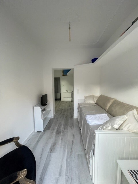 A bright living area features a light-colored pull-out sofa with neatly arranged linens beside a small white TV stand. Light wood flooring complements the white walls. An opening leads to a connected space, with a hint of cabinetry visible in the background.