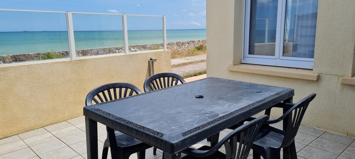 An outdoor dining area features a large table surrounded by four black chairs. The ocean is visible in the background, with calm waters and a clear sky. Sunlight reflects off the table's surface, highlighting its textured design.