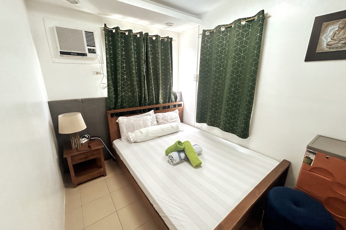 A comfortable sleeping area features a spacious bed with fresh linens and pillows. Green patterned curtains frame a window, allowing natural light to fill the room. A bedside lamp and a small table are positioned beside the bed, complemented by a colorful storage unit.