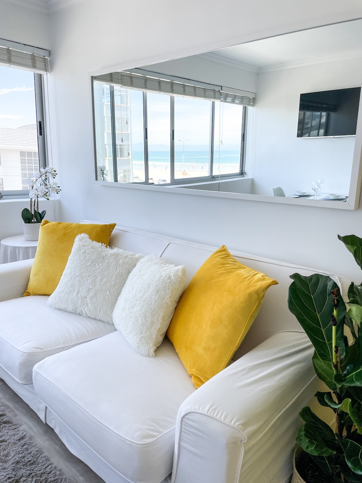 A bright living area features a white sofa adorned with two yellow and one white cushion. A mirror reflects the view of large windows that offer glimpses of the beach and ocean, enhancing the sense of space and natural light.