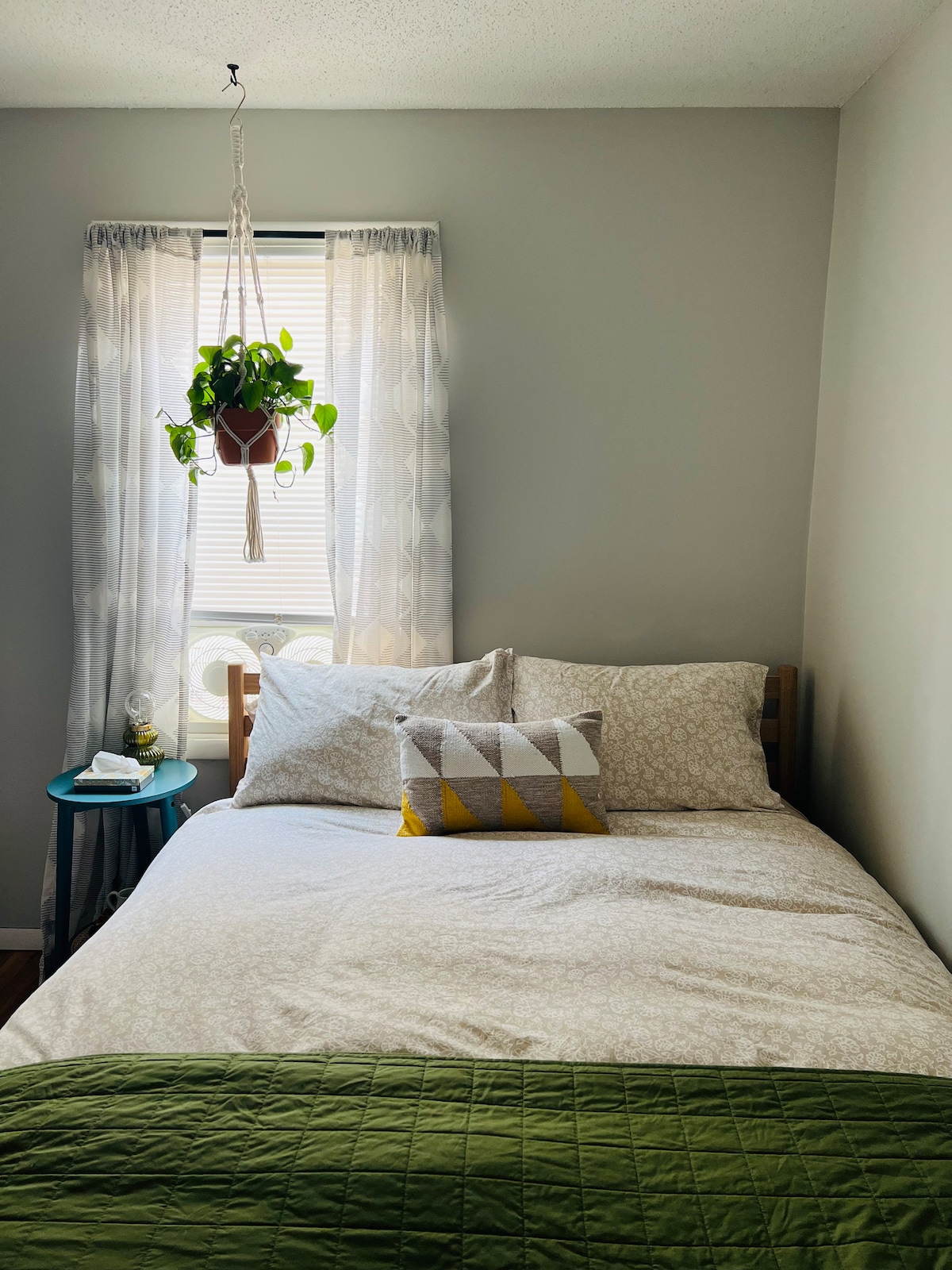 A cozy bedroom features a spacious bed adorned with light-patterned bedding and a yellow accent pillow. A small table beside the bed holds a water carafe. Natural light filters through sheer curtains, and a hanging plant adds a touch of greenery.
