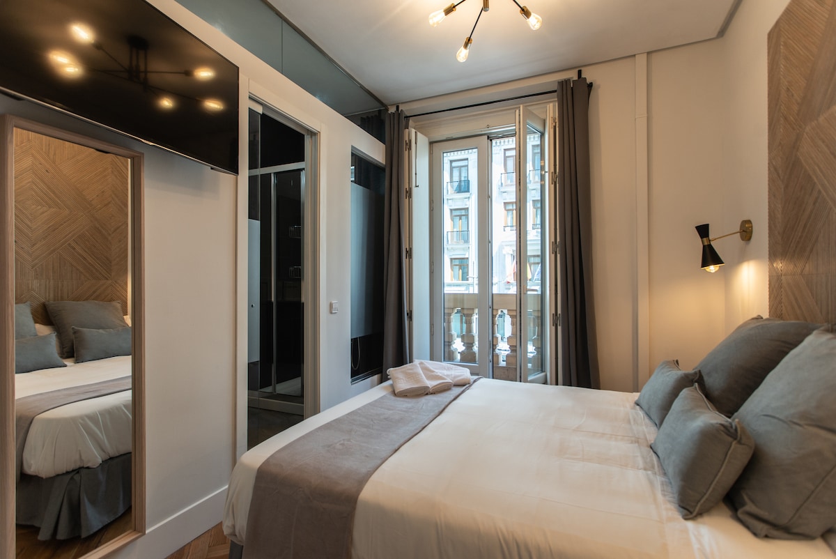 A cozy bedroom features a double bed with soft grey and white linens. A large mirror reflects natural light from the balcony doors, which open to a view of the bustling Gran Vía. Wall sconces provide gentle illumination, enhancing the room's modern design.