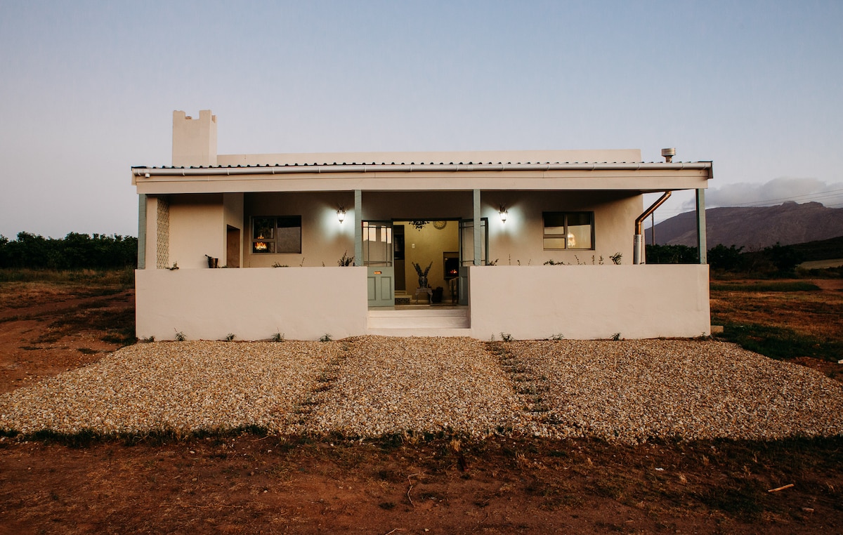 A single-story farmhouse is presented, featuring a welcoming front porch with a covered area. The structure showcases a minimalist design with a light-colored exterior and a gravel pathway leading to the entrance, set against a backdrop of open fields and distant mountains.