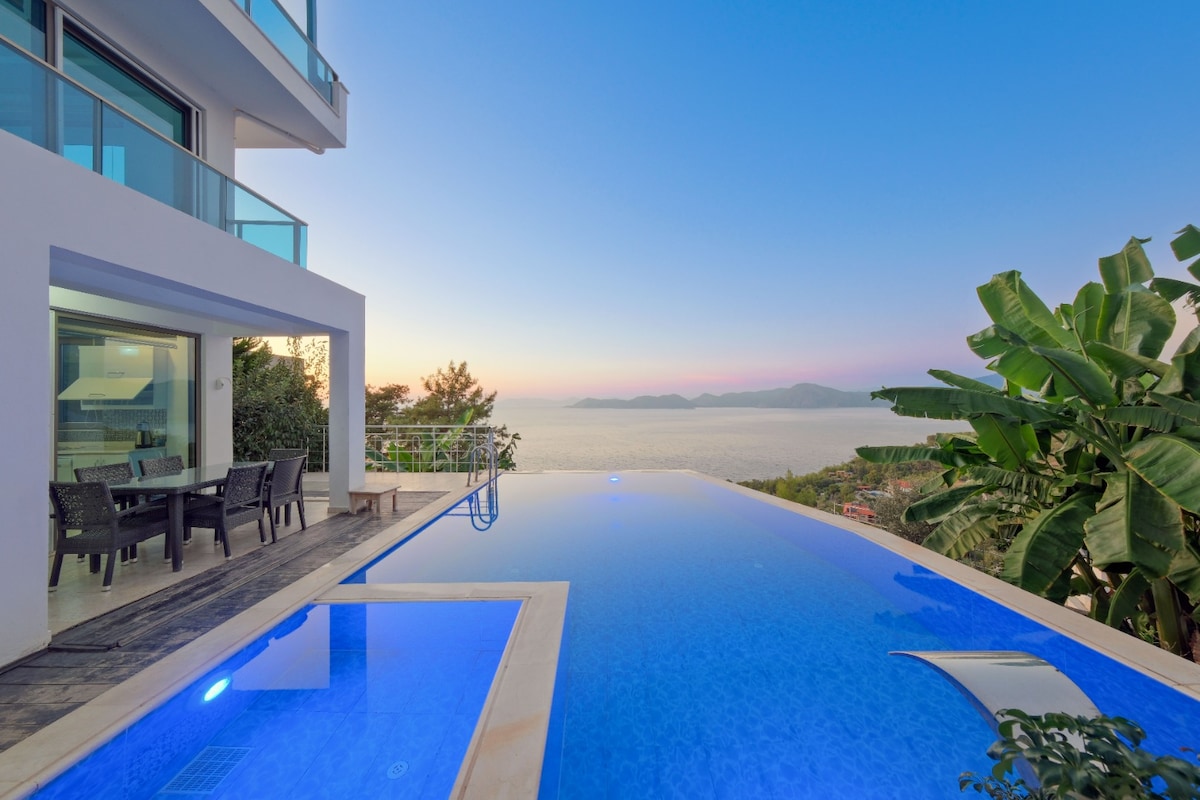 An infinity pool is seen merging with the horizon, reflecting the sunset hues over the ocean. A dining area with dark wicker furniture is positioned nearby, surrounded by green foliage. The modern structure features large glass panels, offering panoramic views of the landscape.