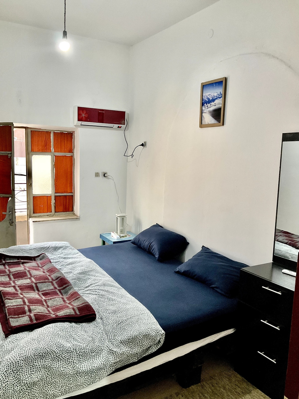 A private bedroom is equipped with a comfortable bed featuring dark blue linens and two pillows. Natural light enters through a window, complemented by wooden shutters. A minimalist bedside table and a dresser provide storage. A framed image adorns the wall, adding a personal touch.
