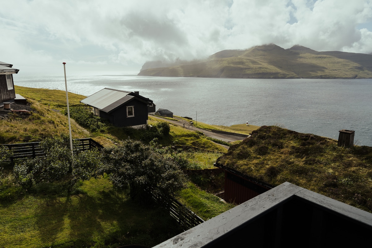 A scenic view captures the tranquil coastline and hills of Leynar, showcasing a blend of green grass and calm waters. The foreground features a rustic house with a green roof, while a dirt road winds along the shoreline, leading to distant hills under a cloudy sky.