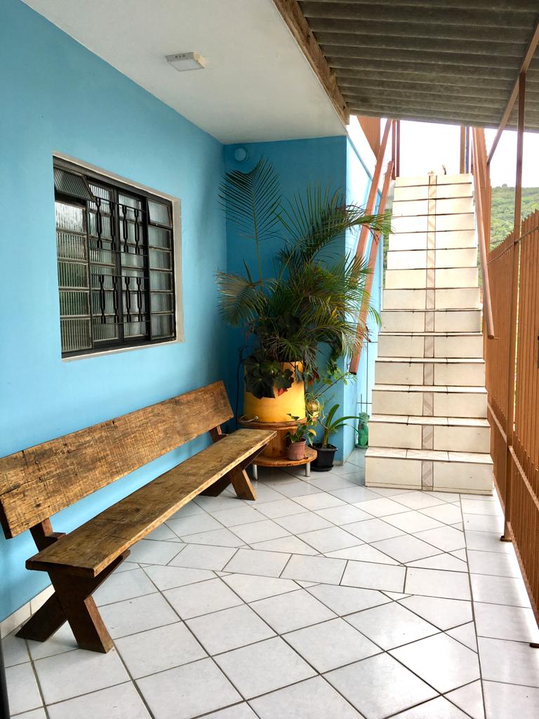 A corridor is illuminated by soft natural light, featuring a wooden bench against a bright blue wall. A potted plant adds a touch of greenery, while tiled flooring leads towards a staircase with white steps, contrasting against the warm tones of the surrounding elements.