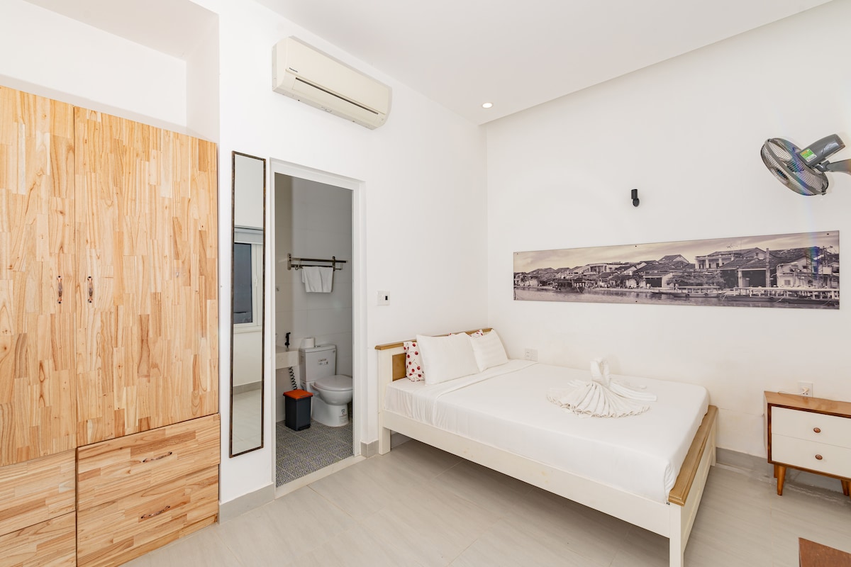 A bright and spacious room features a large bed with a neatly arranged towel display. A wooden wardrobe provides storage, and a mirror is positioned above a vanity space. The room is complemented by a subtle wall decoration and an air conditioning unit on the ceiling.