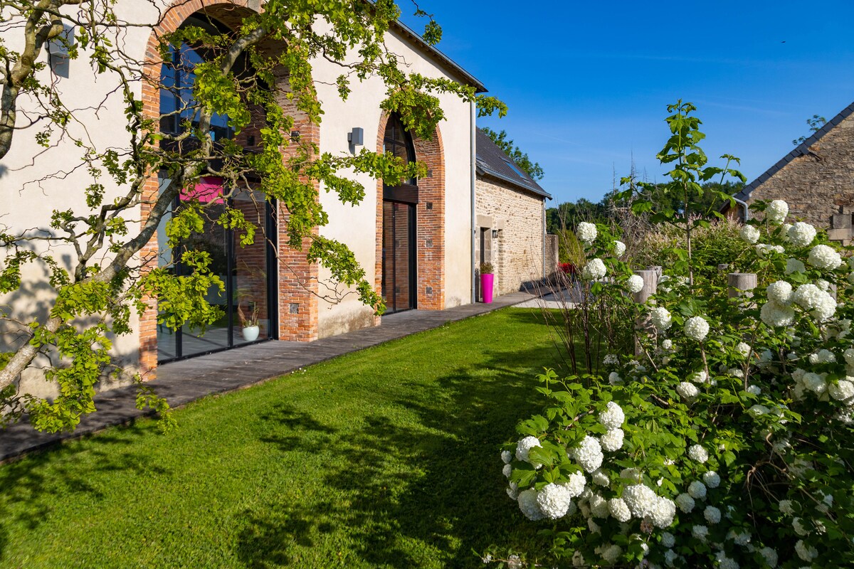 The image presents a charming exterior view of the house, showcasing its architectural details like large arched windows. A well-kept lawn is visible alongside blooming white flowers, enhancing the outdoor space's natural appeal. The clear sky offers a bright backdrop to the setting.