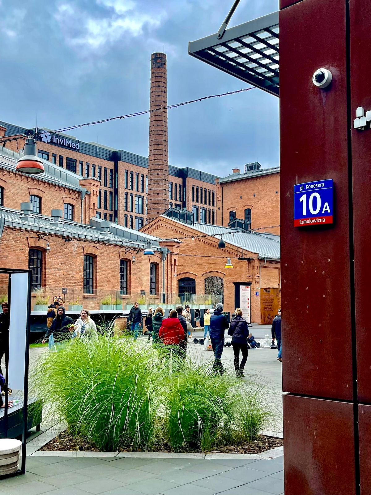 A courtyard is visible, featuring a mix of red brick structures and modern buildings. Green grass and plants add a natural touch to the space. People can be seen walking and gathering in the area, with a prominent chimney rising from one of the historic buildings.