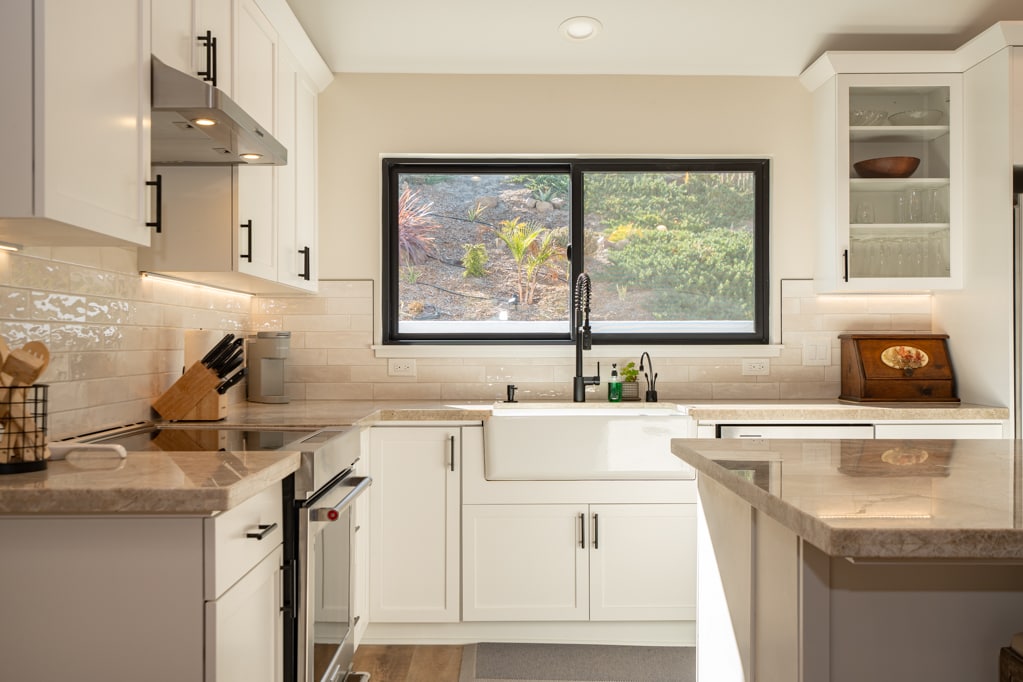 A modern kitchen features sleek white cabinetry and a spacious island with a complementary countertop. Natural light pours in through a large window over a farmhouse sink, highlighting the organized utensils and kitchen tools on the countertops.