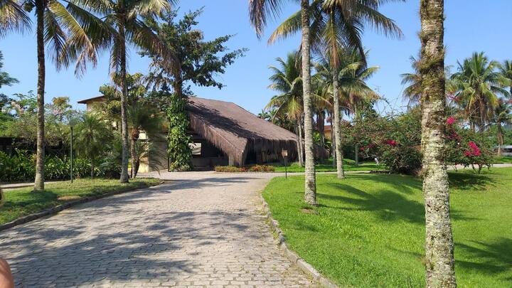A pathway lined with palm trees leads to a resort building featuring a thatched roof. Lush greenery and flowering shrubs are visible on either side, creating a serene entrance to the property under clear blue skies.