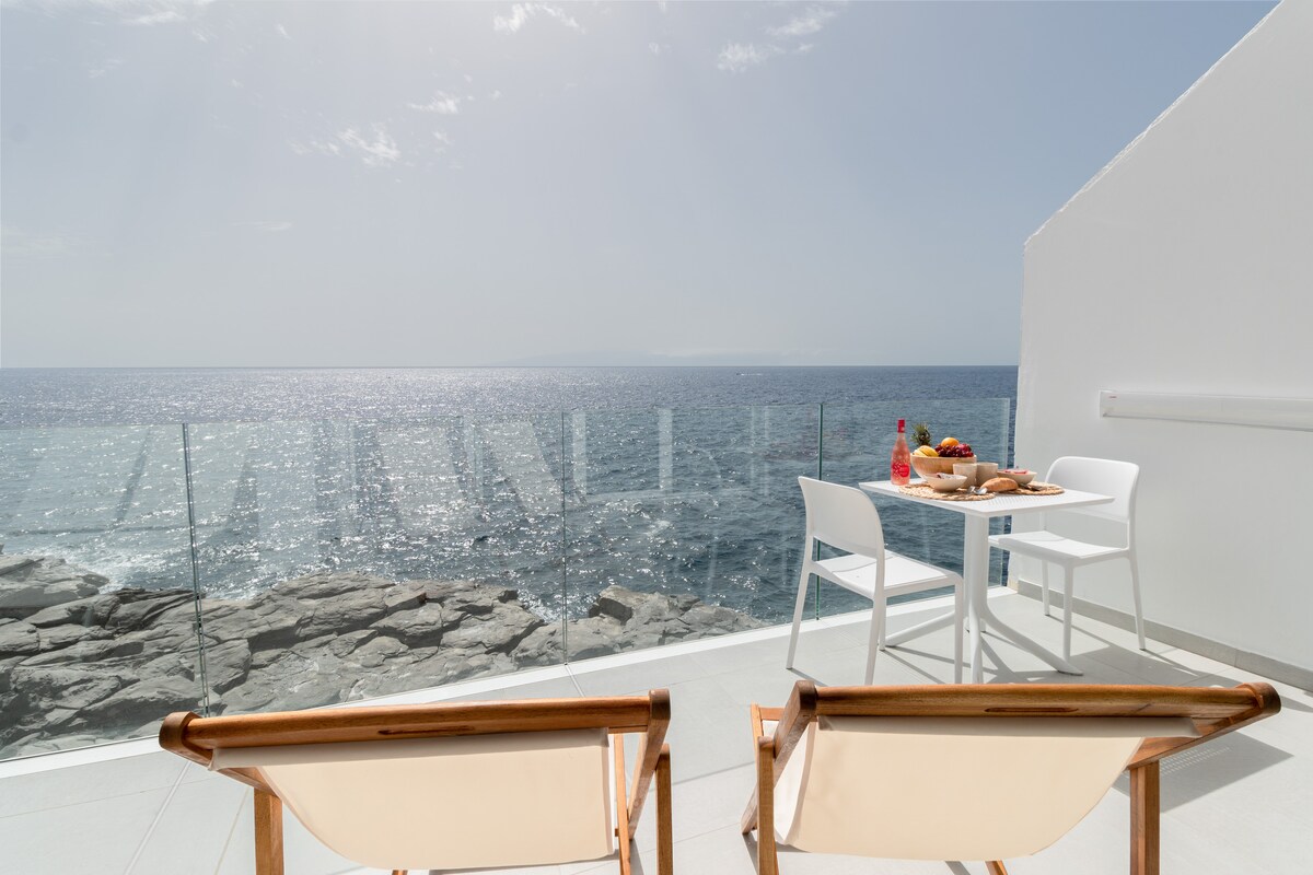 A terrace space is presented with a scenic sea view, featuring a small dining table set for two. Two wooden chairs are placed nearby, offering a comfortable seating area. The sunlight reflects off the water, creating a bright and airy ambiance.