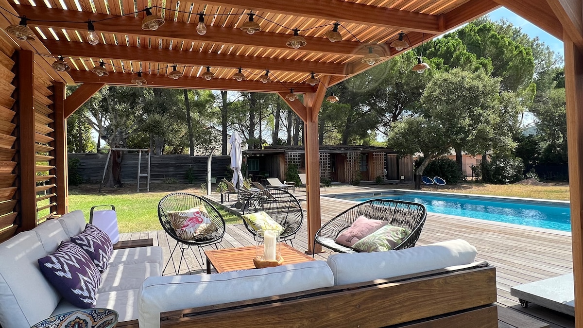 A shaded terrace features a wooden pergola with slatted roof and comfortable seating. Thick cushions adorn the lounge area, accompanied by a wooden table. In the background, a clear swimming pool reflects the surrounding greenery, while sun loungers are positioned nearby.
