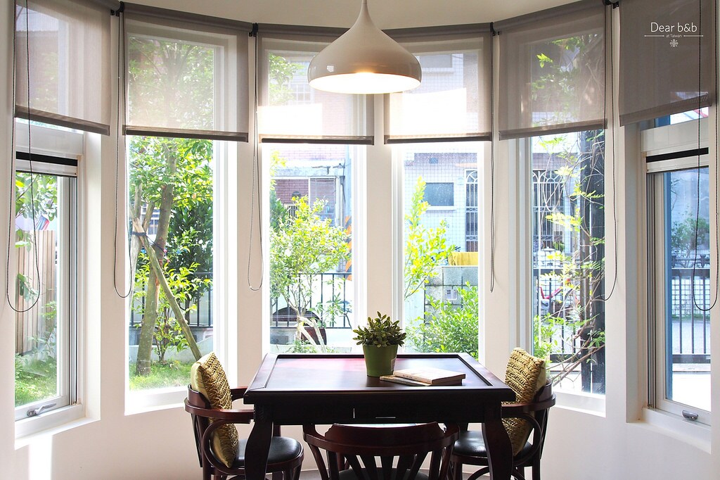 A sunlit dining area is surrounded by large windows that provide ample natural light. A round table is centered in the room, accompanied by four wooden chairs. Greenery can be seen through the windows, creating a refreshing atmosphere.