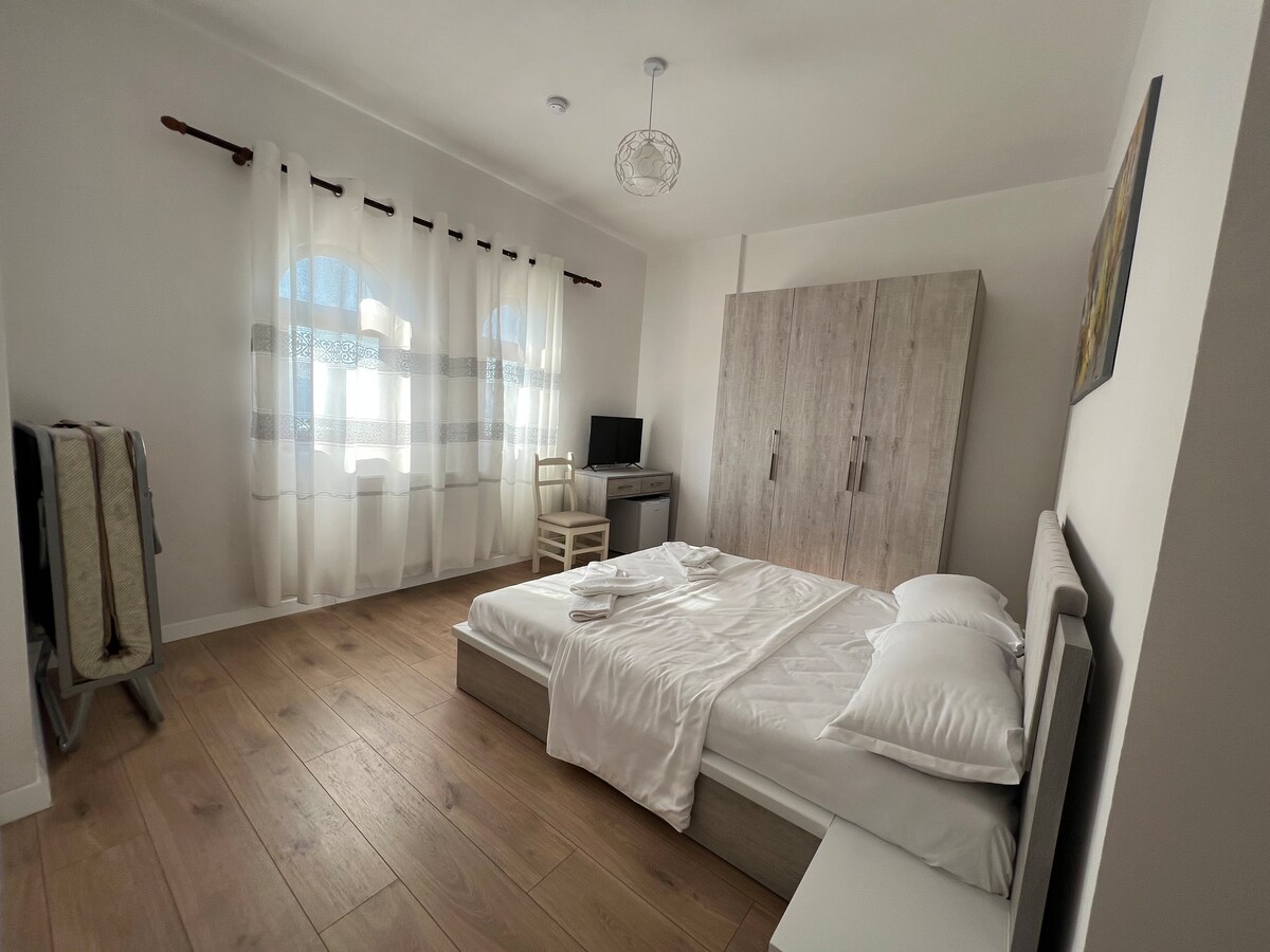 A bright and airy room features a spacious bed with crisp white linens. Natural light filters through sheer curtains adorning large windows. A wooden wardrobe and a small desk with a chair are present, creating a functional yet comfortable space.