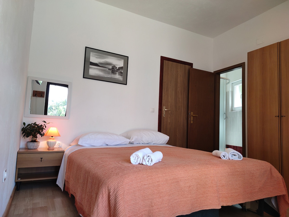 A bedroom features a spacious bed with an orange blanket neatly arranged. Two towels are placed on the bed. A small bedside table with a lamp and a mirror is visible. Light enters through a nearby door leading to a bathroom.