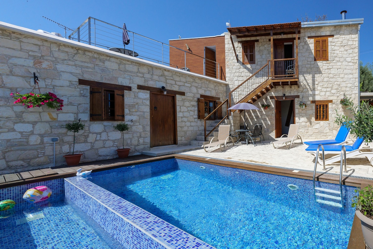 A stone house with a rustic exterior features a private pool surrounded by sun loungers and a shaded umbrella. An outdoor space includes a staircase leading to an upper level, while potted plants add greenery against the stone walls.