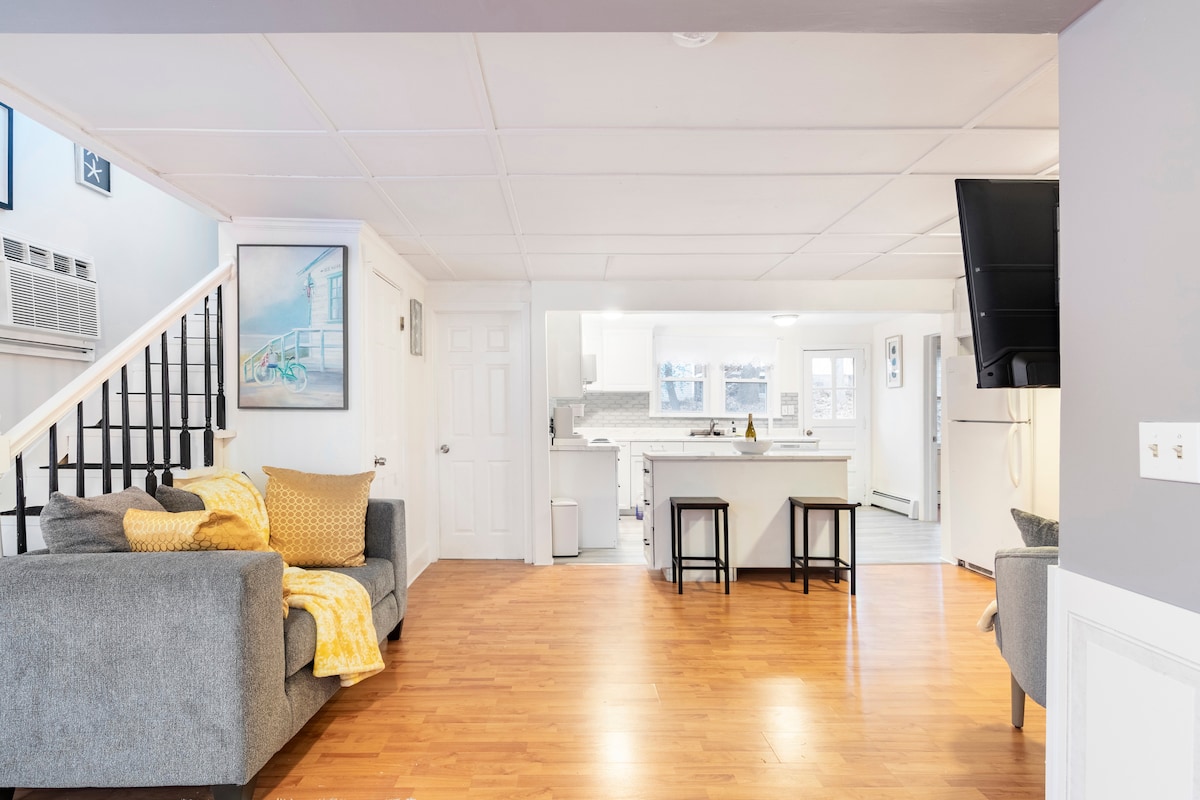 An open living area features a gray sofa adorned with yellow throw pillows. A kitchen with light-colored cabinetry and a large island is visible in the background. Natural light floods the space through multiple windows, enhancing the inviting atmosphere.