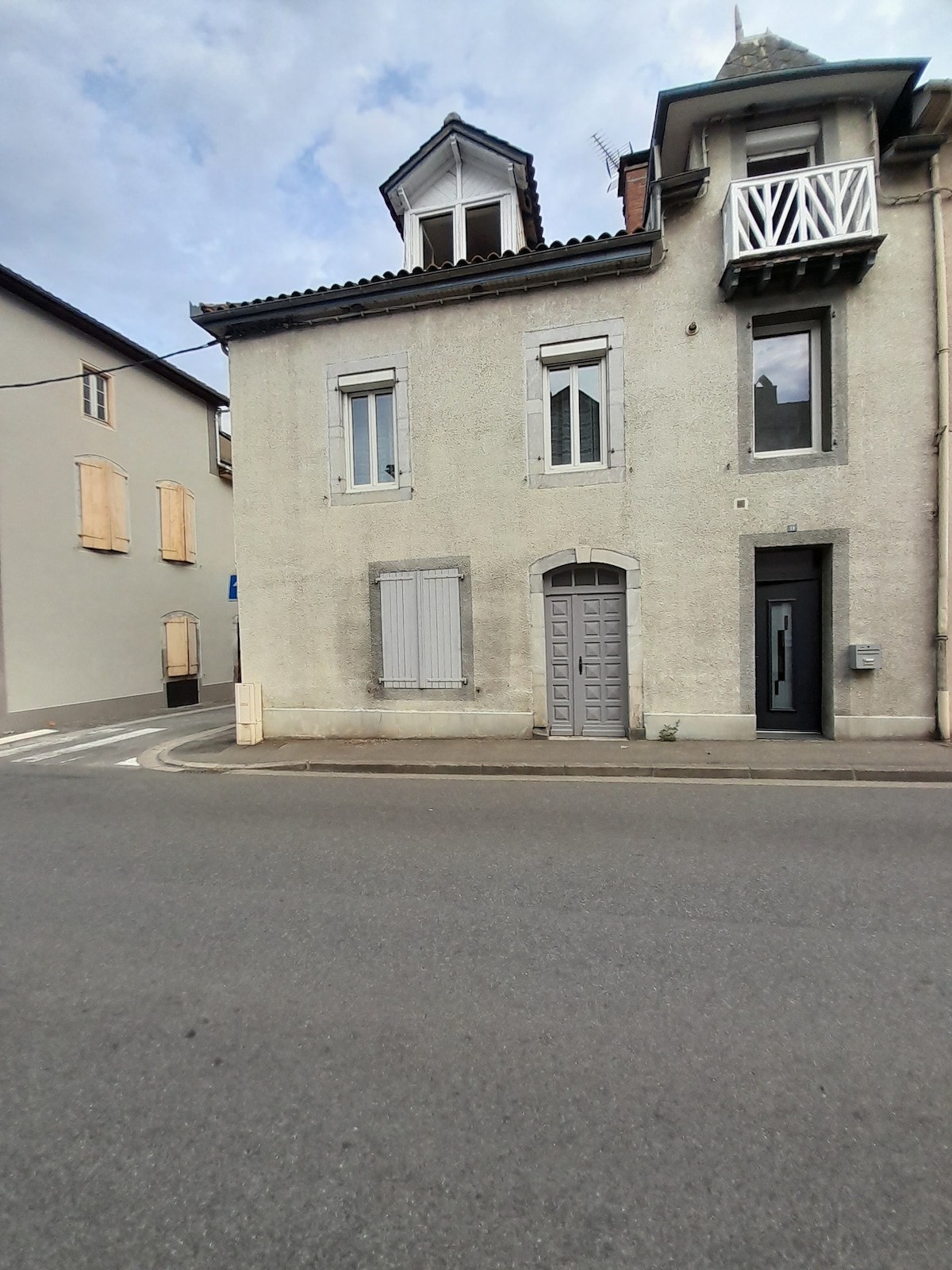 The building features a neutral-colored façade with shutters and a small balcony on the upper level. The front door is framed by modest details, and the surrounding area provides a quiet street view. Windows are present on both levels, enhancing natural light.