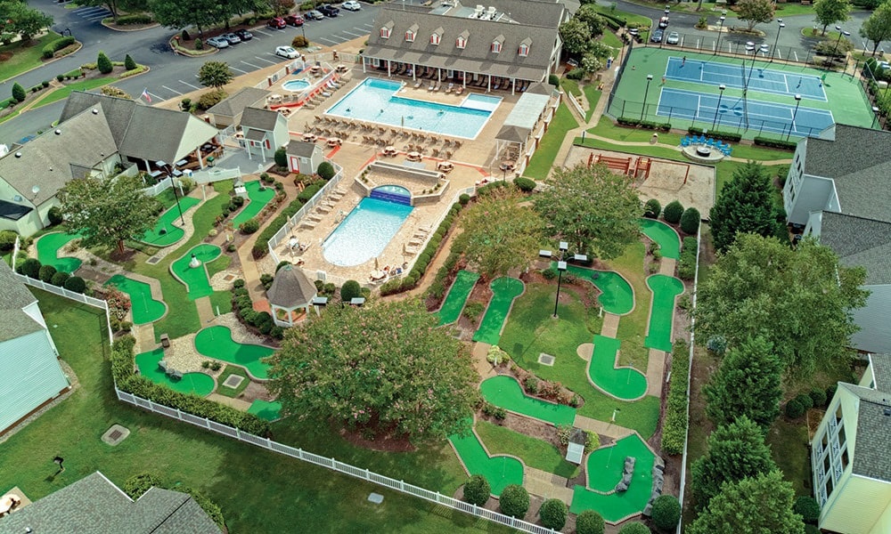 Aerial view showcases a resort-style area featuring a large swimming pool surrounded by lounge chairs. Well-maintained mini-golf courses are visible alongside a tennis court, all framed by lush greenery and neatly arranged landscaping.