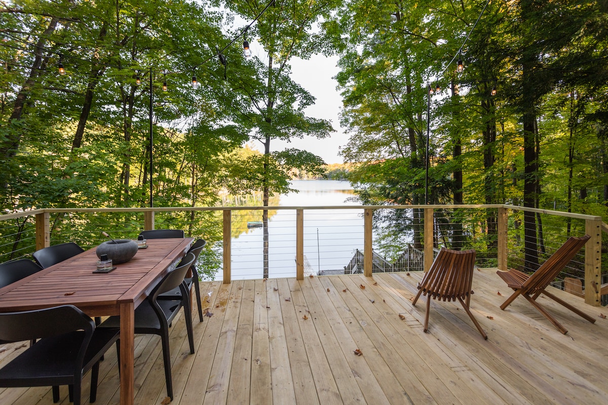 A spacious wooden deck provides an inviting space for outdoor dining and relaxation, adorned with a large dining table and comfortable seating. Lush green trees frame the view, which overlooks the calm lake waters, creating a serene atmosphere perfect for gatherings.