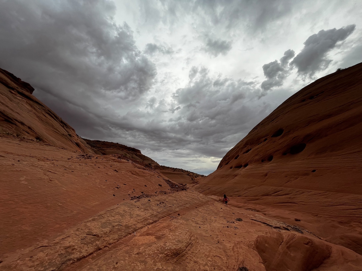 A wide canyon landscape showcases expansive red rock formations under a moody sky filled with clouds. The ground is slightly rugged, leading to a distant figure, providing a sense of scale against the natural beauty of the surroundings.