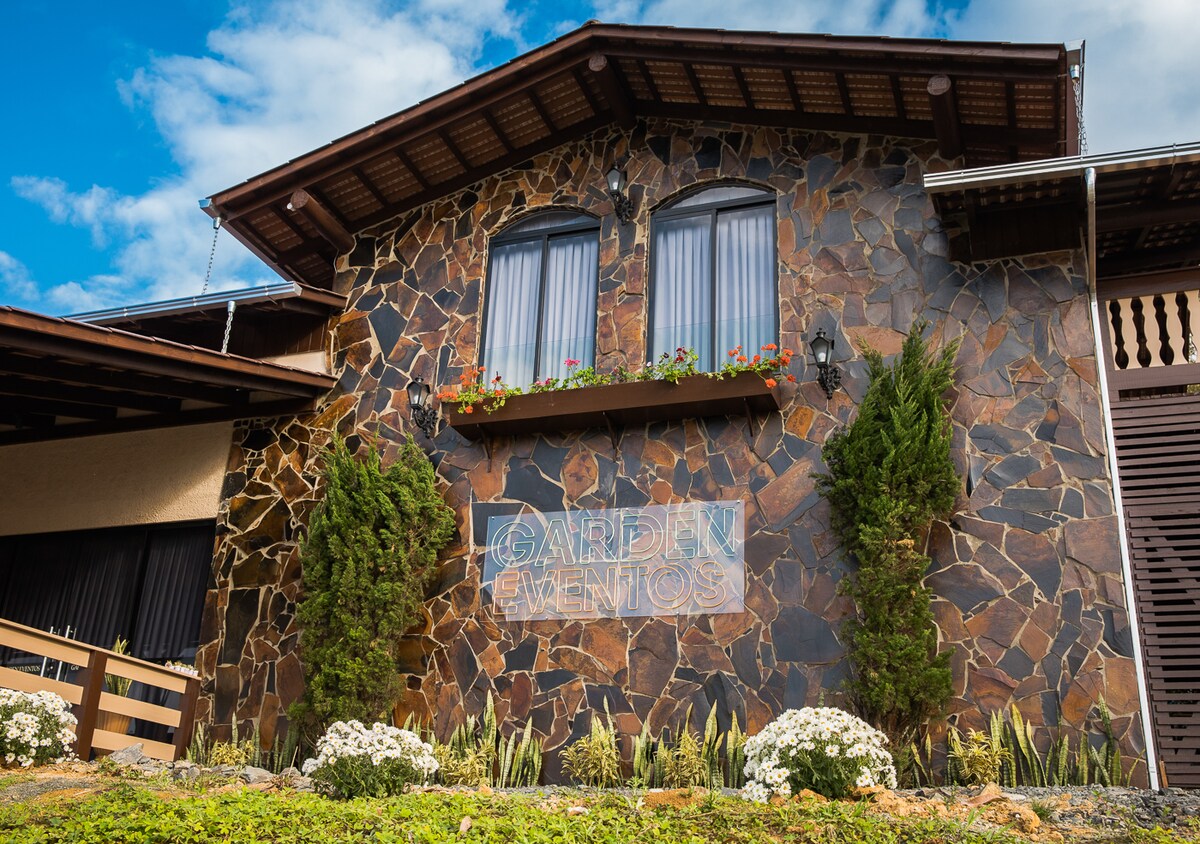 The exterior of a stone structure is showcased, featuring large windows adorned with flower boxes. The signage for 'Garden Events' is displayed prominently. Surrounding greenery includes neatly trimmed shrubs and white flowers, contributing to a welcoming appearance.