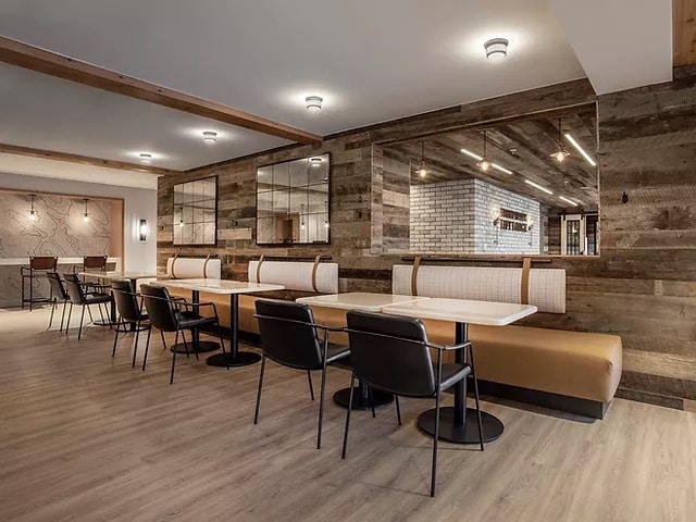 A spacious dining area is characterized by light wood flooring and rustic wooden walls. There are multiple booths with cushioned seating and sleek black chairs arranged around white tables. Large mirrors enhance the sense of space, reflecting the warm lighting throughout the area.