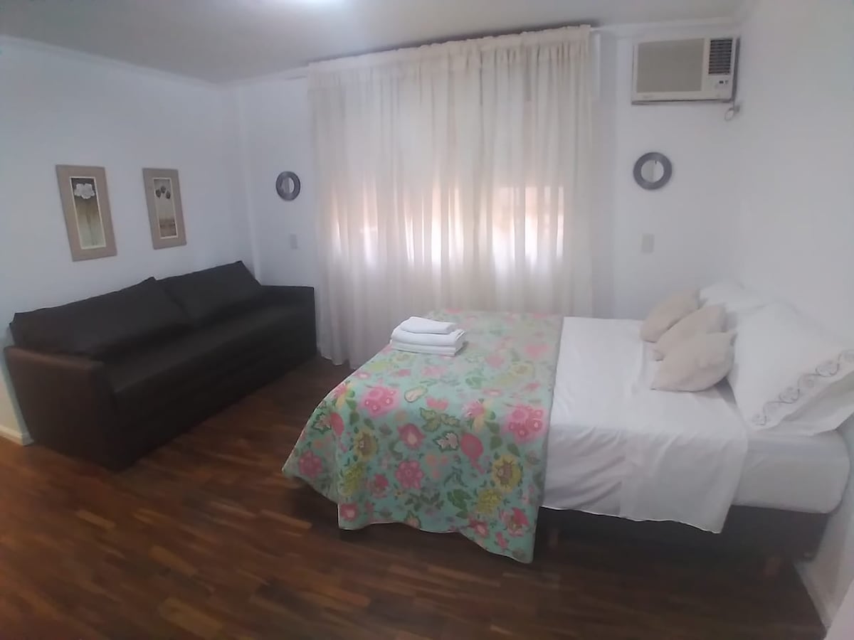 A bright sleeping area features a spacious bed draped in a floral blanket, accompanied by two decorative pillows. A dark sofa is positioned nearby, and soft light filters through sheer curtains, enhancing the cozy ambiance. Wall art and mirrors add subtle touches to the room.