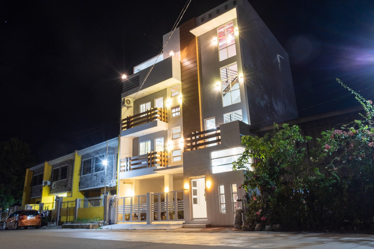 A modern multi-story building is illuminated at night, featuring spacious balconies with wooden railings. Bright lights accentuate the clean lines of the architecture. Surrounding greenery adds a natural touch, while vehicles parked nearby reflect the lively atmosphere of the neighborhood.