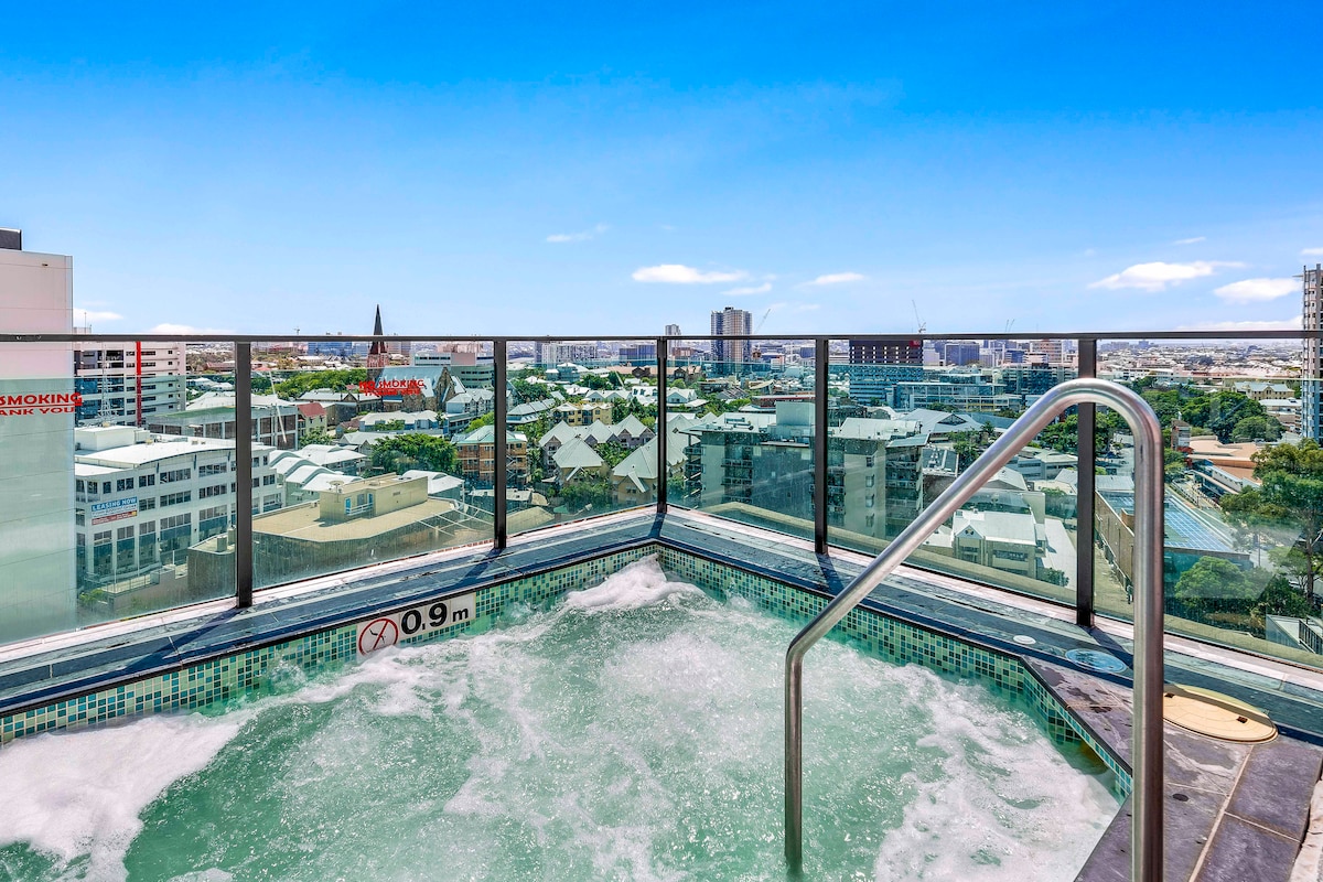 A rooftop area showcases a bubbling hot tub, with clear views of the surrounding cityscape. Glass safety barriers provide an unobstructed sight of the skyline, while the sunlit scene enhances the relaxing atmosphere.