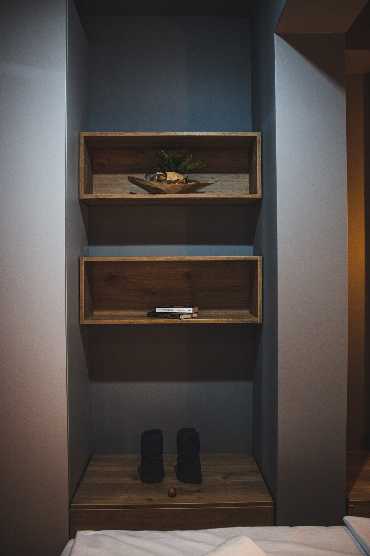 A built-in shelving unit is mounted against the wall, featuring two wooden shelves with decorative items, including a small plant. The base includes a drawer that showcases a pair of rolled towels, contributing to the organized and minimalist aesthetic of the space.