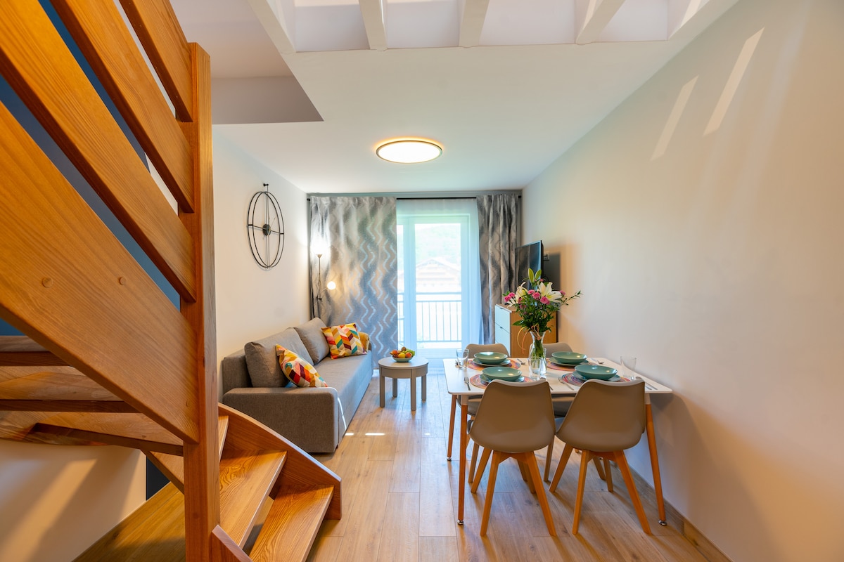A contemporary dining area is visible, featuring a table set for six. A comfortable sofa adorned with colorful cushions complements the space. Natural light enters through a glass door leading to a balcony, while wooden stairs add an inviting element to the interior.