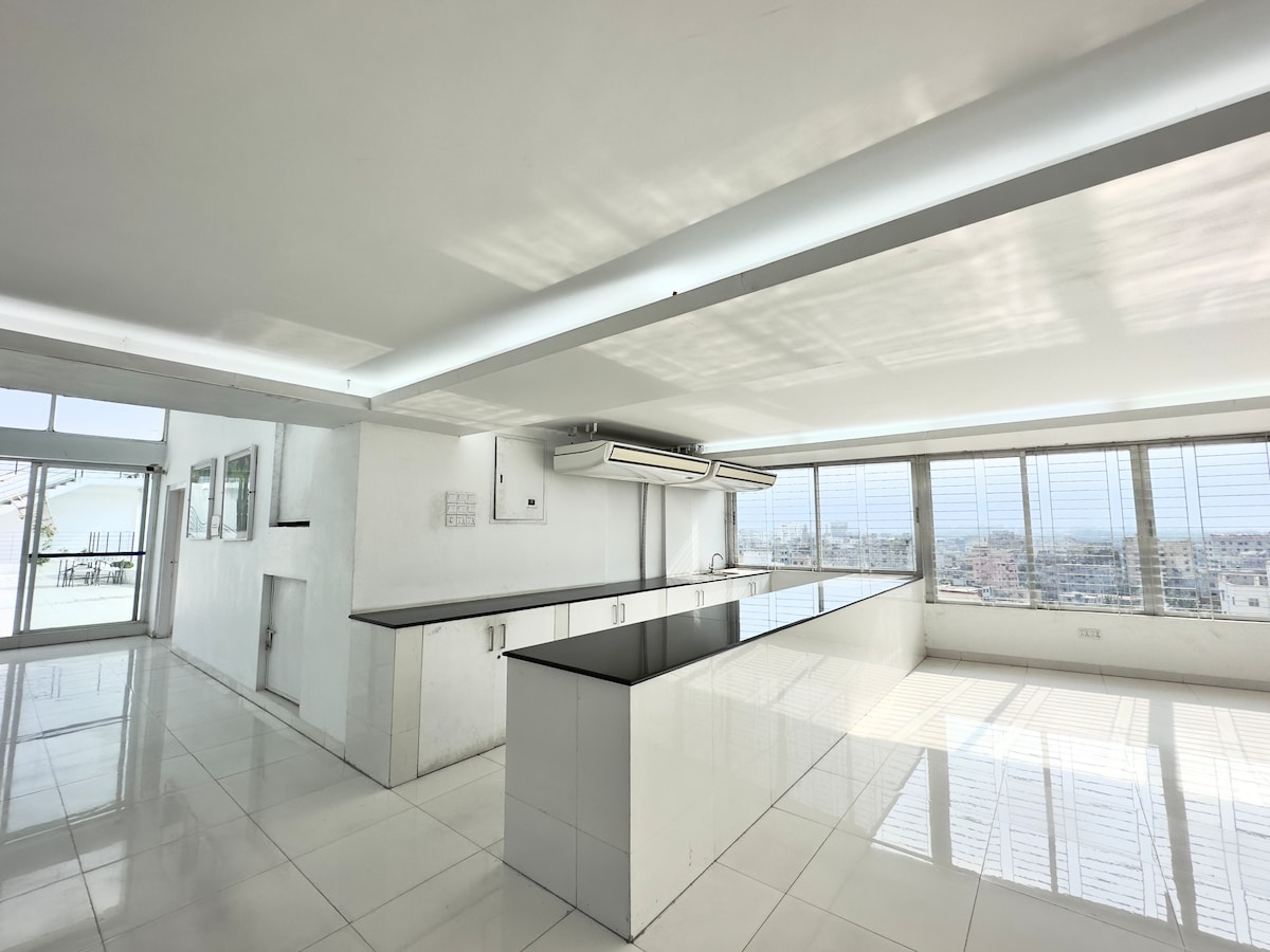 A spacious studio apartment is showcased with white tile flooring and ample natural light. The open kitchen features a large countertop and seating area. Large windows provide a view of the surroundings, while the minimalist design enhances the sense of openness.