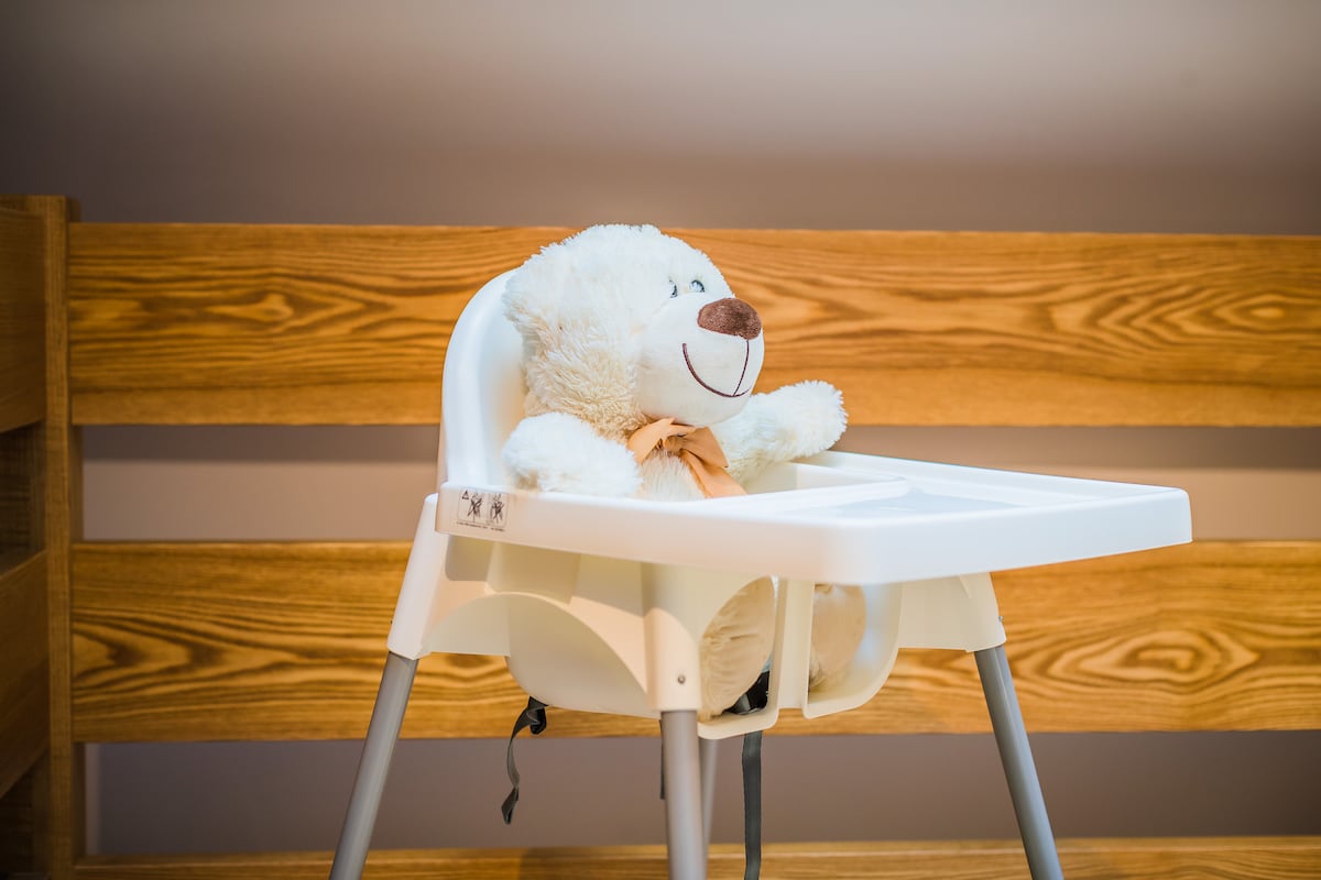 A high chair is positioned against a wooden backdrop, featuring a friendly stuffed bear seated in it. The chair is designed for young children, equipped with a simple safety harness and a smooth, easy-to-clean surface.