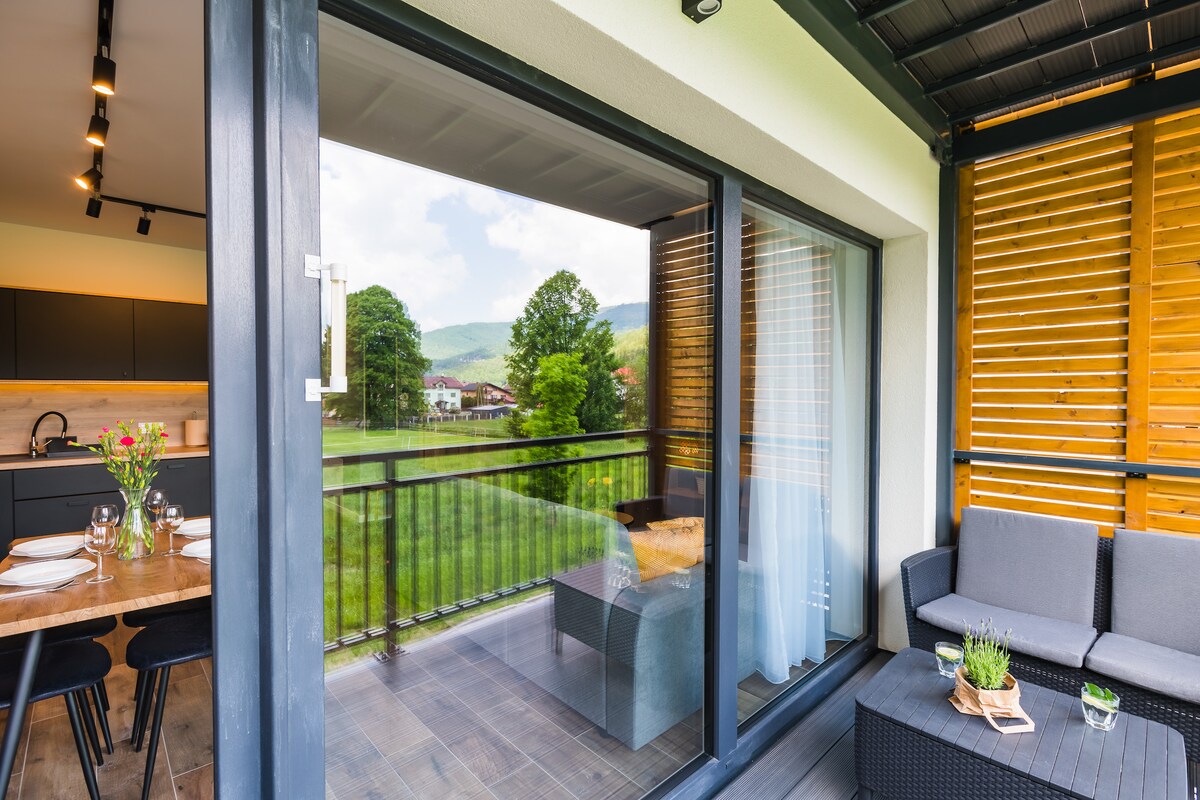 A balcony area is displayed with modern outdoor seating arrangements. Large glass doors open to reveal a view of greenery and distant mountains. Natural light brightens the space, with wooden slats providing privacy and a sense of connection to the outdoors.