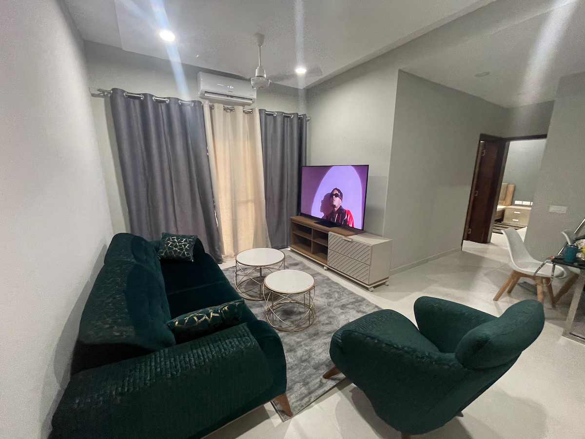 A comfortable living area is highlighted by a plush green sofa and a matching armchair. Two round coffee tables are placed on a soft area rug. Large windows dressed with sheer and dark curtains allow natural light, while a television sits on a modern media console.