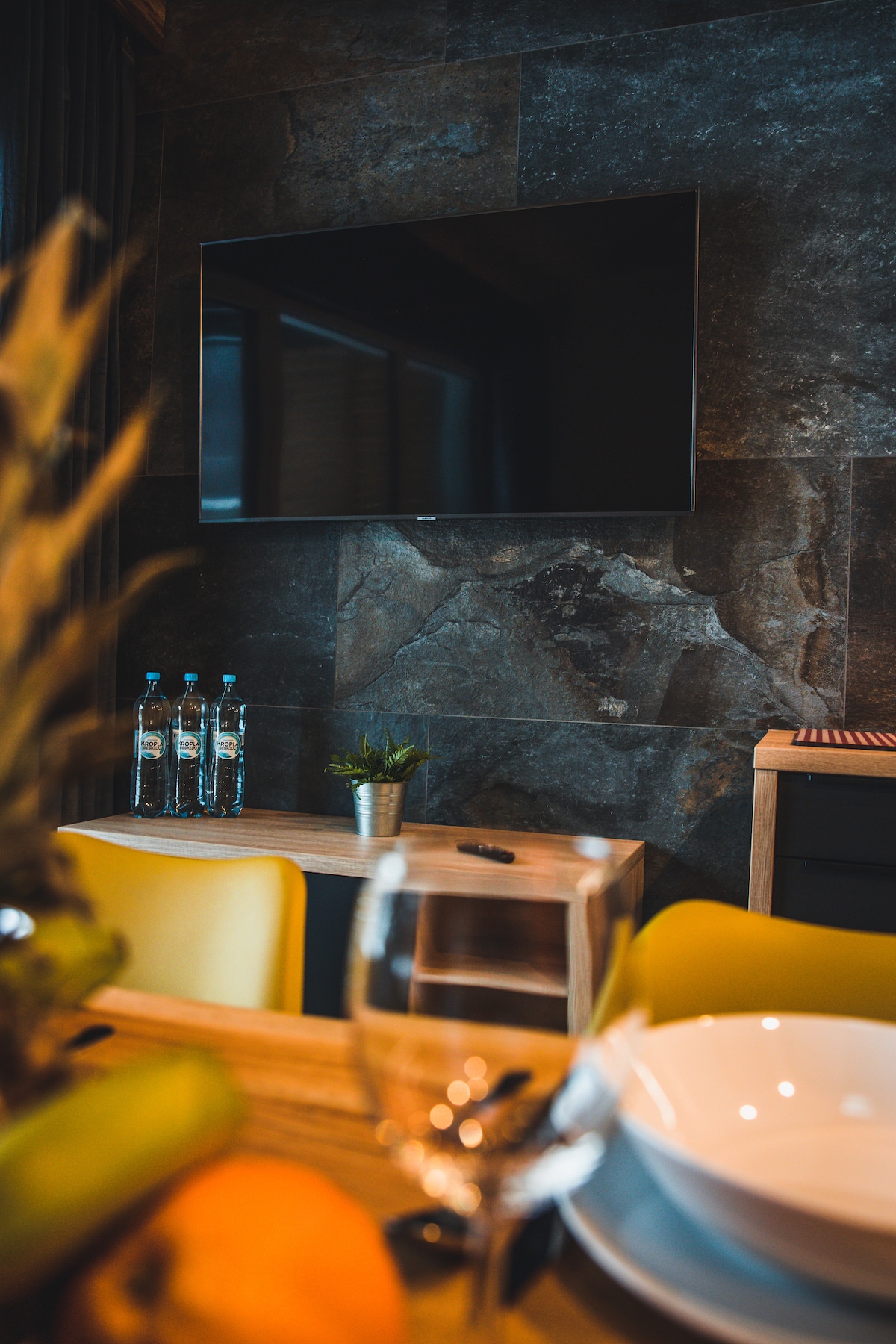 A modern living area features a sleek flat-screen television mounted on a textured stone wall. Below the TV, a simple wooden console is complemented by a small potted plant. Nearby, a dining table is set with glassware and dishes, enhancing the inviting space.