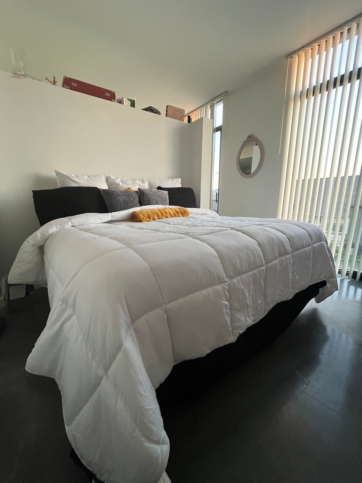 A spacious bed is dressed in a white comforter, with multiple decorative pillows arranged neatly. A round mirror is mounted on the wall beside the bed, and sunlight filters through vertical blinds, creating soft highlights on the interior walls.