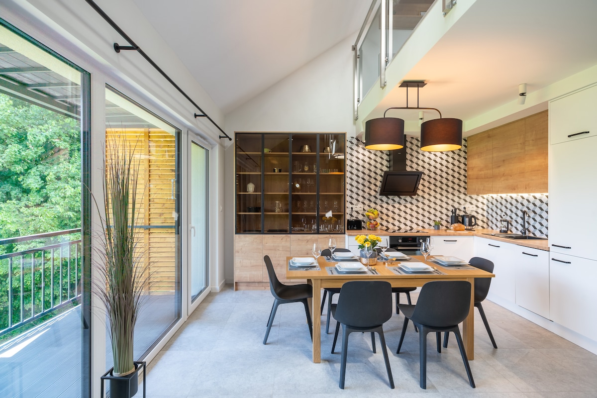 A modern kitchen and dining area are fitted with sleek cabinetry and a large wooden dining table. Four black chairs surround the table, while a geometric backsplash adds visual interest. Large windows provide natural light and views of greenery outside.