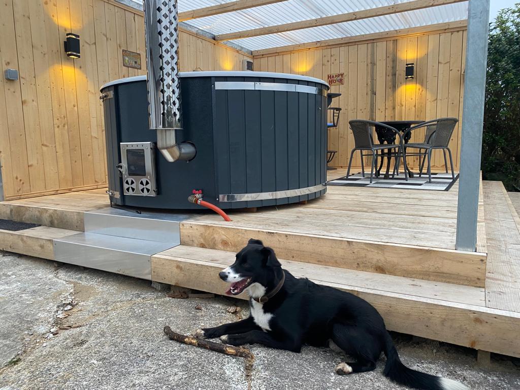 An outdoor area features a wooden deck with a circular hot tub beside a chimney. Comfortable seating is arranged nearby, with a black dog resting on the ground. Soft lighting is mounted on the wooden walls, creating an inviting ambiance.