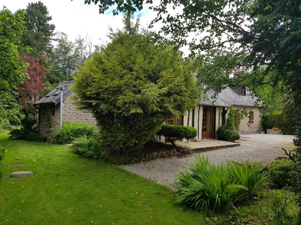 The exterior of the house is set within a lush garden, featuring a variety of greenery and a gravel path leading to the entrance. A large, well-maintained tree and shrubs provide natural privacy, while the stone facade of the house complements the surroundings.