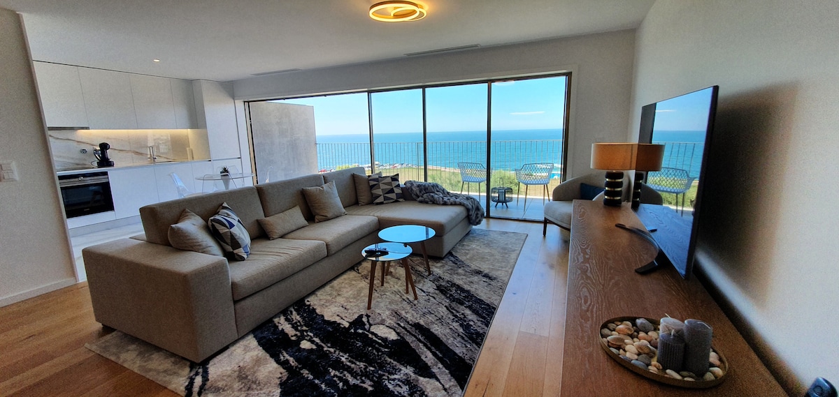 The spacious living area features a large sectional sofa, a round coffee table, and a decorative area rug. Floor-to-ceiling windows provide views of the ocean, and natural light fills the room. A modern television is positioned on a wooden console, enhancing the contemporary décor.