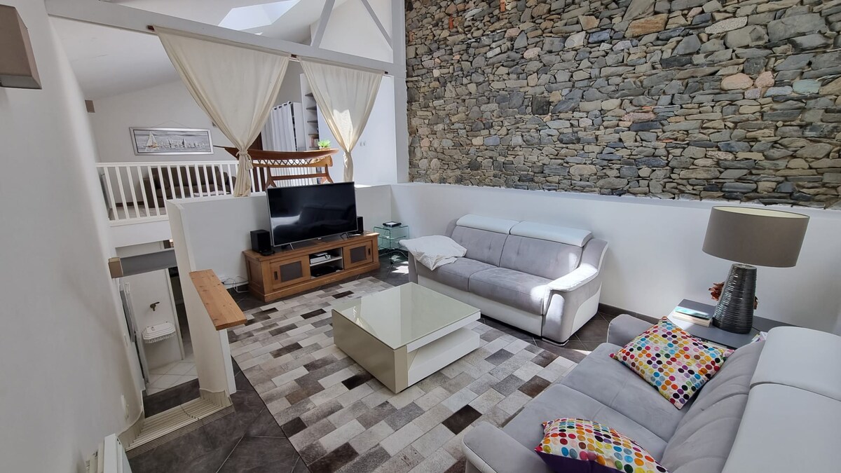 The spacious living area features a stone accent wall, a comfortable sofa, and a television on a wooden console. Natural light filters through the high ceilings and large windows, creating a bright atmosphere. A patterned rug adds texture to the floor.