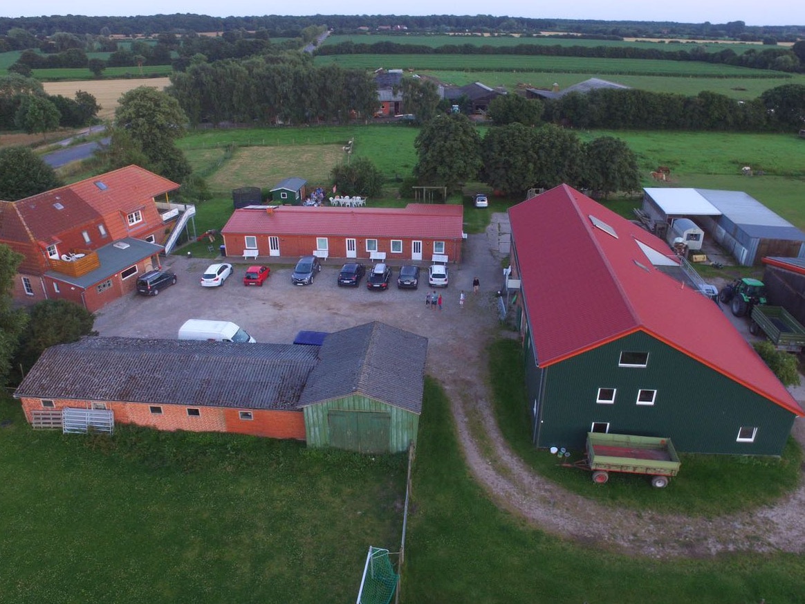 An aerial view of a spacious farm complex is displayed, featuring several buildings with red and green roofs. A parking area is visible with multiple vehicles. Surrounding fields and trees contribute to the serene rural setting.
