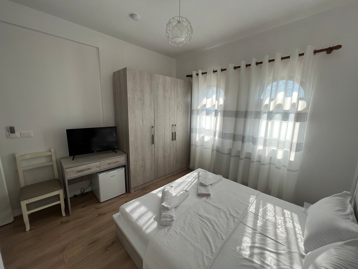 A bright bedroom features a spacious bed with neatly arranged linens. Natural light filters through sheer curtains over two windows. A compact wardrobe stands beside a television and a small refrigerator, complemented by a chair and a minimalist desk.